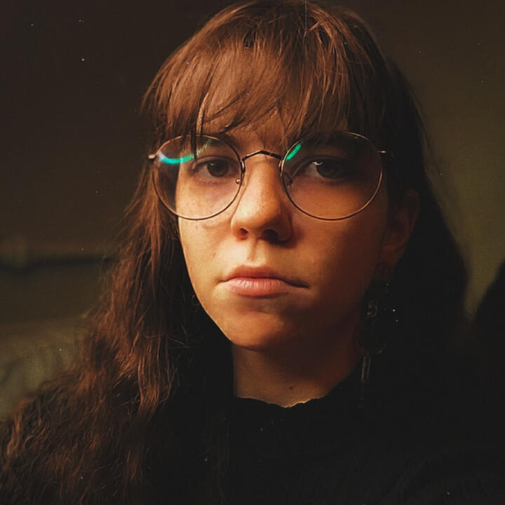 image of a white nonbinary individual with long brown hair, bangs, and round glasses